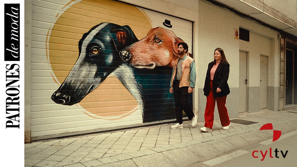 Foto portada del proyecto para Patrones de Moda donde se ve a un hombre y una mujer caminando por una acera delante de un local cuya verja es un dibujo de dos perros galgo